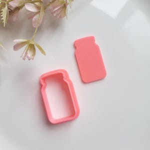 May include: Two pink plastic cookie cutters shaped like mason jars. One cutter is solid and the other is an outline.