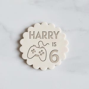 May include: A light beige, scalloped-edge cookie with the text "HARRY IS 6" and a controller graphic. The design is etched into the cookie's surface. A fun, celebratory treat. The cookie is a perfect birthday cake topper.