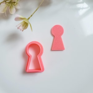 May include: Two pink plastic keyhole cookie cutters. The cutters are in the shape of a keyhole, with a rounded top and a rectangular bottom.