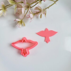 May include: Two pink plastic cookie cutters shaped like birds. The cutters are on a white surface.