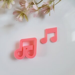 May include: Two pink plastic musical note cookie cutters. The larger cutter is a double note and the smaller cutter is a single note.