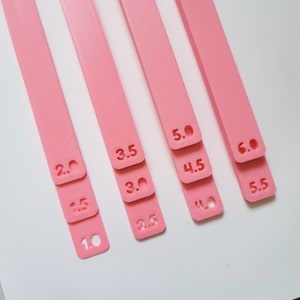 May include: Four pink plastic rulers with different measurements marked on them. The rulers are stacked on top of each other, with the largest ruler at the bottom and the smallest ruler at the top. The measurements on the rulers are 1.0, 1.5, 2.0, 2.5, 3.0, 3.5, 4.0, 4.5, 5.0, 5.5, and 6.0.