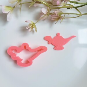 Pode incluir: Dois cortadores de biscoitos de plástico rosa em forma de lâmpada mágica de Aladim. O cortador maior é um contorno detalhado da lâmpada, enquanto o cortador menor é uma forma sólida.