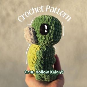Puede incluir: Un peluche de ganchillo Grub Hollow Knight verde y amarillo hecho a mano, con un ojo negro y un hocico beige. El texto "Crochet Pattern" y "Grub Hollow Knight" está en la imagen.