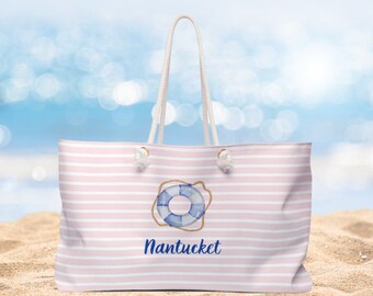 May include: A large beach tote bag with pink and white horizontal stripes. The bag features a blue and white life preserver graphic and the word "Nantucket" in blue script. The bag has white rope handles and is sitting on sand.