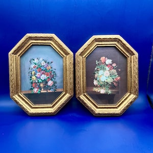 May include: Two framed floral still life paintings. Each painting features a bouquet of colorful flowers in a vase. The frames are gold-colored with an octagonal shape. The background of one painting is blue, and the other is brown.