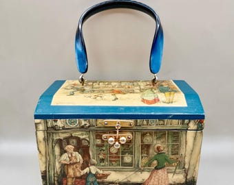 May include: A rectangular box-style handbag with a dark blue arched handle and a gold-tone metal clasp. The exterior features a detailed illustration of a clockmaker's shop with figures, a dog, and clocks, while the lid shows a street scene.
