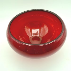May include: A vibrant, translucent red glass bowl with a smooth, rounded silhouette and a wide, open top. The bowl features a deep, polished interior and a thick, curved rim, showcasing a consistent, bold crimson color throughout the glass structure.
