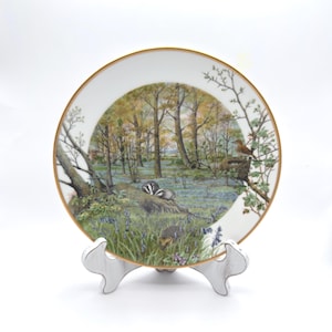 May include: A decorative plate with a gold rim, featuring a detailed woodland scene. The artwork depicts a forest with bluebells, trees, and wildlife, including badgers and a fox. The plate is displayed on a white stand.
