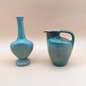 May include: Two ceramic vases in a teal glaze. One is a tall, slender vase with a rounded base. The other is a pitcher with a handle and a leaf design. Both are on a light beige background.