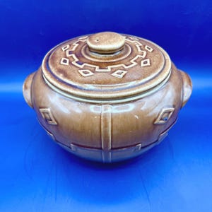 May include: A brown ceramic casserole dish with a lid. The dish has a round shape with two handles and geometric designs. The lid has a small knob and a similar geometric pattern. The dish is a warm brown color.