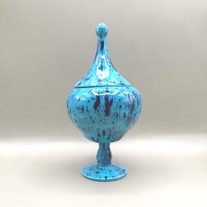 May include: A turquoise ceramic jar with a lid, featuring a speckled pattern of dark blue spots. The jar has a bulbous body, a pedestal base, and a tall, slender finial on the lid. The overall height is approximately 12 inches.