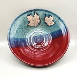 May include: A handmade ceramic bowl with a tri-color design. The top section is teal, transitioning to blue, and then to a deep red at the base. Two copper-colored leaf accents are placed on the teal section. The bowl has a glossy finish.