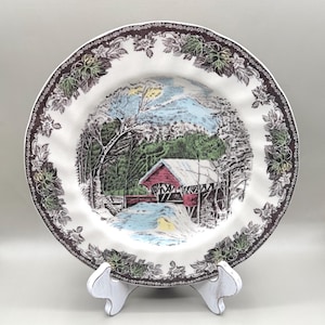 May include: A round ceramic plate featuring a central illustration of a red covered bridge in a snowy landscape with trees and a stream. The rim is decorated with a dark brown border of intricate leaf and floral patterns on a white background.
