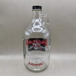 May include: Clear glass half-gallon jug with a black cap and handle. The jug features a red and black graphic with the words "Graveyard of the Atlantic" and "Atlantic" printed on the front. The jug is empty.