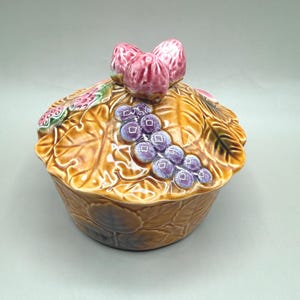 May include: A ceramic lidded serving dish with a leaf and grape design. The dish is brown with purple grapes and pink accents on the lid. The lid is topped with three pink fig-like shapes.