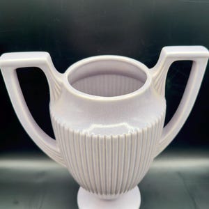 May include: A light purple ceramic vase with two handles and a pedestal base. The vase has vertical ribbed detailing around the body. The handles are rectangular and extend outward from the rim.