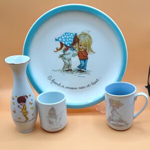 May include: A set of four vintage porcelain pieces featuring a girl with blonde hair and a girl with brown hair wearing a blue and white polka dot hat. The pieces include a plate with a blue rim, a vase, a mug, and a small cup. The plate has the text "A friend is someone near at heart". The vase has the text "Good morning sunshine". The mug has the text "Love". The cup has the text "Be Happy".