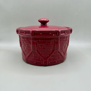 May include: A red ceramic casserole dish with a lid. The dish has a basket weave design and a small knob on the lid. The casserole dish is a deep red color and appears to be made of ceramic.