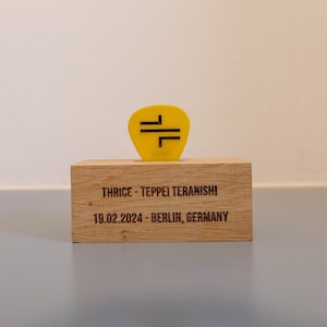 Può includere: Un plettro per chitarra giallo con un design nero è esposto su un blocco di legno. Il blocco ha inciso il testo "THRICE - TEPPEI TERANISHI 19.02.2024 - BERLINO, GERMANIA".
