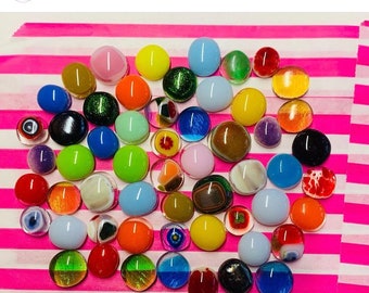 Puede incluir: Una colección de piezas redondas de vidrio fundido de varios colores. Las piezas de vidrio están dispuestas en forma de corazón sobre una superficie rayada de color rosa y blanco. Las piezas de vidrio son de varios colores, incluyendo rojo, naranja, amarillo, verde, azul, morado y marrón.