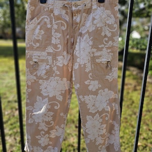 May include: Tan cargo trousers featuring a white floral design. The trousers have two front pockets, two cargo pockets, and a drawstring waist. They are suspended from a black hanger.