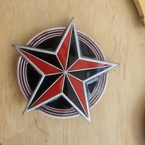 May include: A metal star-shaped emblem with a red and black design. The star is set against a black circular background with concentric rings. The emblem has a silver border and is on a wooden surface.