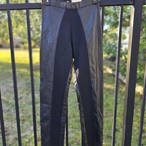 Puede incluir: Pantalones de cuero negro con un panel central de tela negra. Los pantalones cuelgan de una percha negra sobre un fondo borroso al aire libre. Las secciones de cuero están a los lados.