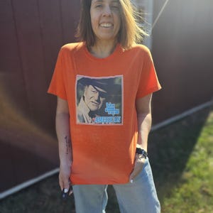 May include: A person wearing an orange t-shirt with a picture of John Wayne on it. The shirt says "American" on the bottom. The person is standing in front of a red barn.