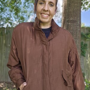 Peut inclure: Veste matelassée marron avec un col montant et deux poches avant. La veste a une fermeture boutonnée et une coupe décontractée. La personne porte une chemise noire sous la veste.