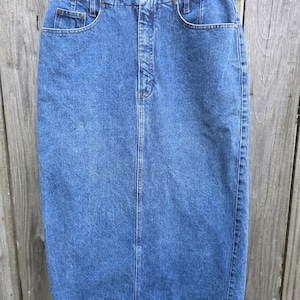May include: A blue denim skirt hanging on a white hanger. The skirt has a classic five-pocket design, a button and zipper closure, and a straight silhouette. The denim is a medium wash with subtle fading.