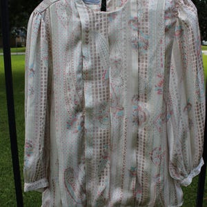 May include: A vintage blouse with a floral and geometric pattern in beige, pink, and blue. The blouse has long sleeves, a round neckline, and the brand name "Notations" on the label.