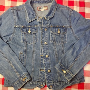 Puede incluir: Una chaqueta vaquera azul con tachuelas plateadas alrededor de los bolsillos y el cuello. La chaqueta tiene cierre de botones y mangas largas.