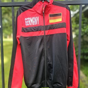 May include: A red, black, and white track jacket with the word "GERMANY" printed across the chest. The jacket features the German flag on the right chest and a full-length zipper. The sleeves and cuffs are red.