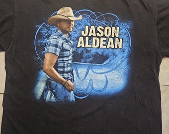 May include: Black t-shirt with a blue and white graphic of country music singer Jason Aldean. The graphic features Aldean wearing a cowboy hat and a plaid shirt, standing in front of a blue and white background with the text "Jason Aldean".
