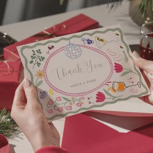 May include: A thank you card with a floral and cocktail-themed design. The card features an oval frame with the words "Thank You" and the names "NADIA + CHAD". The card is held by a person, with a red envelope and gift box in the background.