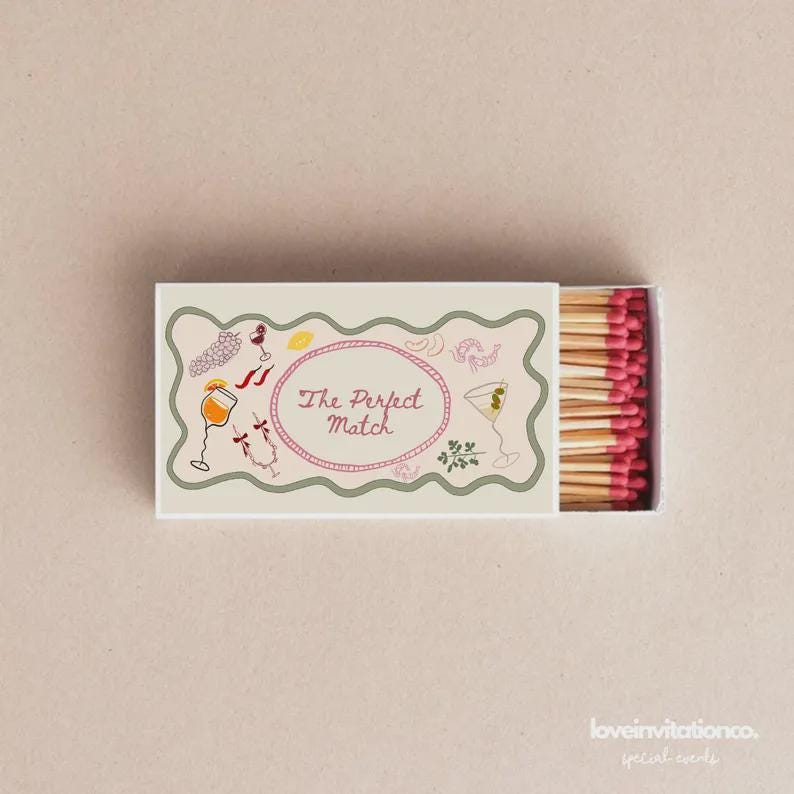May include: A rectangular matchbox with a decorative border and the text "The Perfect Match" inside a pink oval. The box features illustrations of cocktails and food. The matches have pink tips.