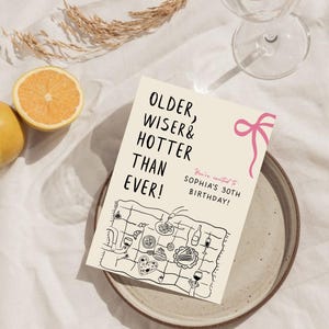 May include: A white card with black text that reads "Older, Wiser & Hotter Than Ever!" and "You're invited to Sophia's 30th Birthday!"  The card has a pink ribbon and a black and white illustration of a picnic blanket with food and drinks.