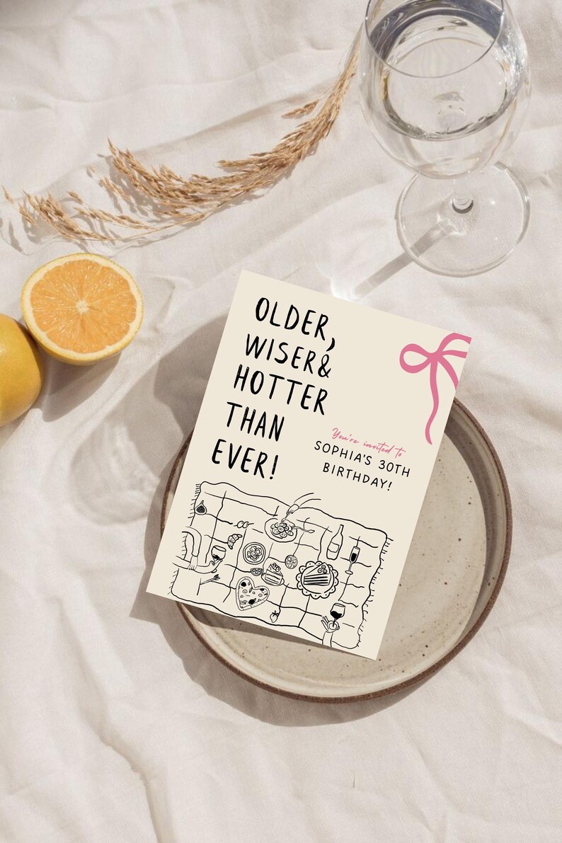 May include: A white card with black text that reads "Older, Wiser & Hotter Than Ever!" and "You're invited to Sophia's 30th Birthday!"  The card has a pink ribbon and a black and white illustration of a picnic blanket with food and drinks.