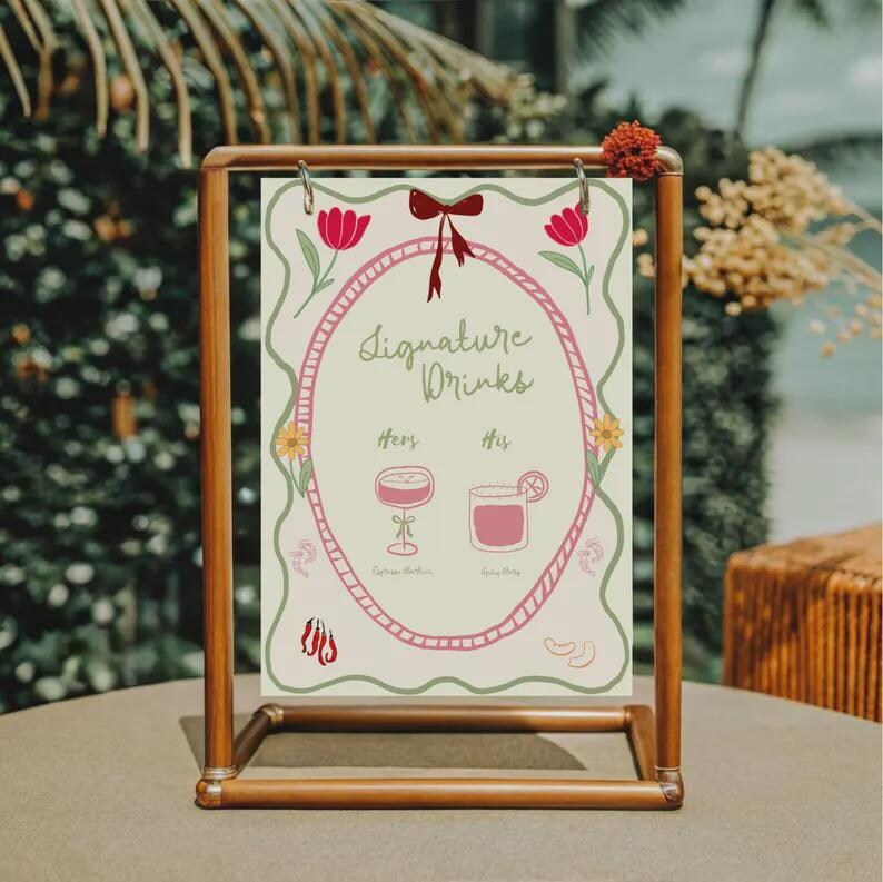 May include: A copper-colored metal frame holds a sign with the words "Signature Drinks." The sign features a pink oval border with floral and bow accents. The sign has illustrations of two drinks, labeled "Hers" and "His."