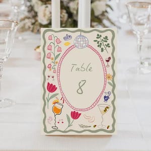 May include: A table number card with a cream background, featuring an oval frame with the word "Table" and the number "8". The card is decorated with illustrations of flowers, cocktails, and a disco ball. The card is on a white tablecloth.