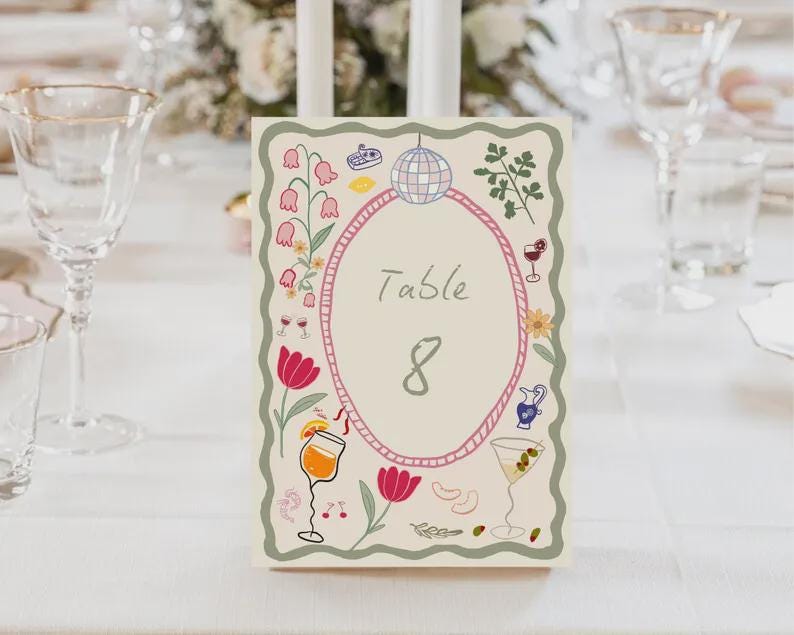 May include: A table number card with a cream background, featuring an oval frame with the word "Table" and the number "8". The card is decorated with illustrations of flowers, cocktails, and a disco ball. The card is on a white tablecloth.