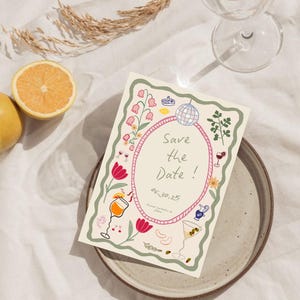 May include: A cream-colored invitation card with a floral border and the text "Save the Date!" in a pink oval. The card features illustrations of cocktails, flowers, and a disco ball. A glass of water and a halved lemon are in the background.