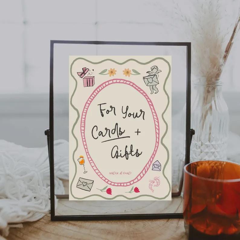 May include: A framed sign with the text "For Your Cards + Gifts" in a decorative oval. The sign has a cream background with illustrations of gifts, flowers, and envelopes. The frame is black, and the sign is displayed on a wooden surface.