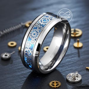 May include: A silver-colored ring with a blue and silver gear design. The ring has a polished finish and the word "Tungsten" is visible on the side. The ring is surrounded by small watch parts.