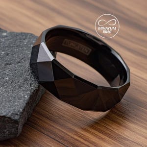 May include: A black tungsten ring with a faceted design. The ring is resting on a dark gray stone and a wooden surface. The word "TUNGSTEN" is engraved on the inside of the ring. The ring has a geometric, modern design.