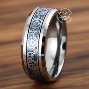 May include: A silver-colored ring with a blue inlay and a gear design. The ring has a polished finish and a detailed pattern of interlocking gears and other mechanical elements. The ring is a close-up shot, showcasing the intricate design.