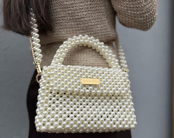 Peut inclure: Un sac à main en perles blanches avec une poignée supérieure dorée et une étiquette en métal doré qui indique "SIMON MILLER".