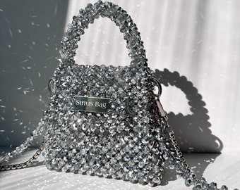 May include: A small, silver beaded handbag with a top handle and a chain shoulder strap. The bag has a rectangular shape and a metal tag that reads "Sirius Bag". The beads are clear and reflective, giving the bag a sparkly appearance.