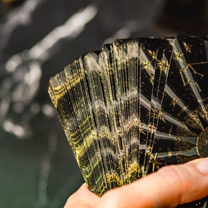 May include: A deck of tarot cards with a black and gold design featuring stars and a sun with a face. The cards are fanned out in a hand.